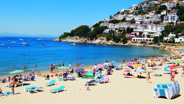 A beach with sun loungers and umbrellas, a clear blue sea, and buildings on a hillside.