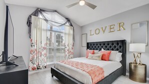 Un dormitorio con cama, televisor, ventilador de techo, ventana con cortinas y una pared con la palabra "LOVER".