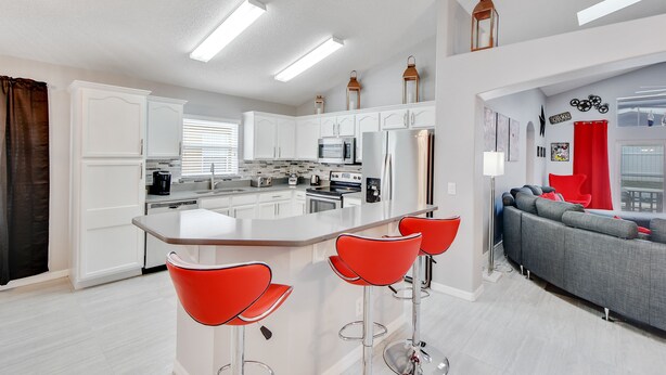 Una cocina moderna con armarios blancos, una isla central con taburetes rojos y un sofá gris en la sala de estar.