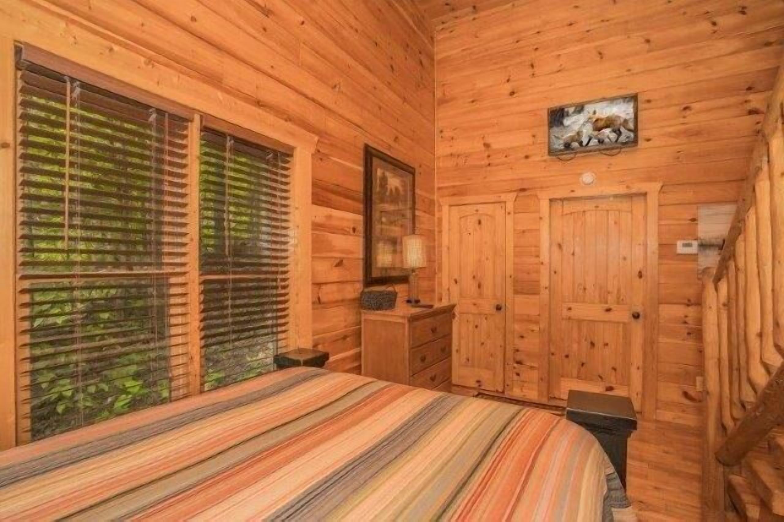 Ein Zimmer einer Holzhütte mit einem Bett, einem Fenster mit Jalousien, einer Tür und einem Bild an der Wand.
