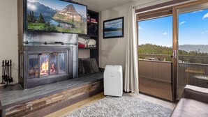 A living room with a fireplace, a television mounted above it, and a balcony with a view of mountains. - Modern Edelweiss 2 bedroom Condo! Fantastic views and some of the best ski access!! (Whitefish)