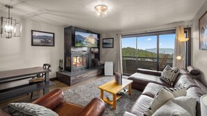 A modern living room with a fireplace, comfortable seating, and large windows offering a scenic view. - Modern Edelweiss 2 bedroom Condo! Fantastic views and some of the best ski access!! (Whitefish)