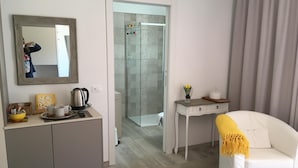 Une chambre d’hôtel avec un lit, un fauteuil blanc, un bureau agrémenté d’un vase de fleurs, un miroir et un espace douche.