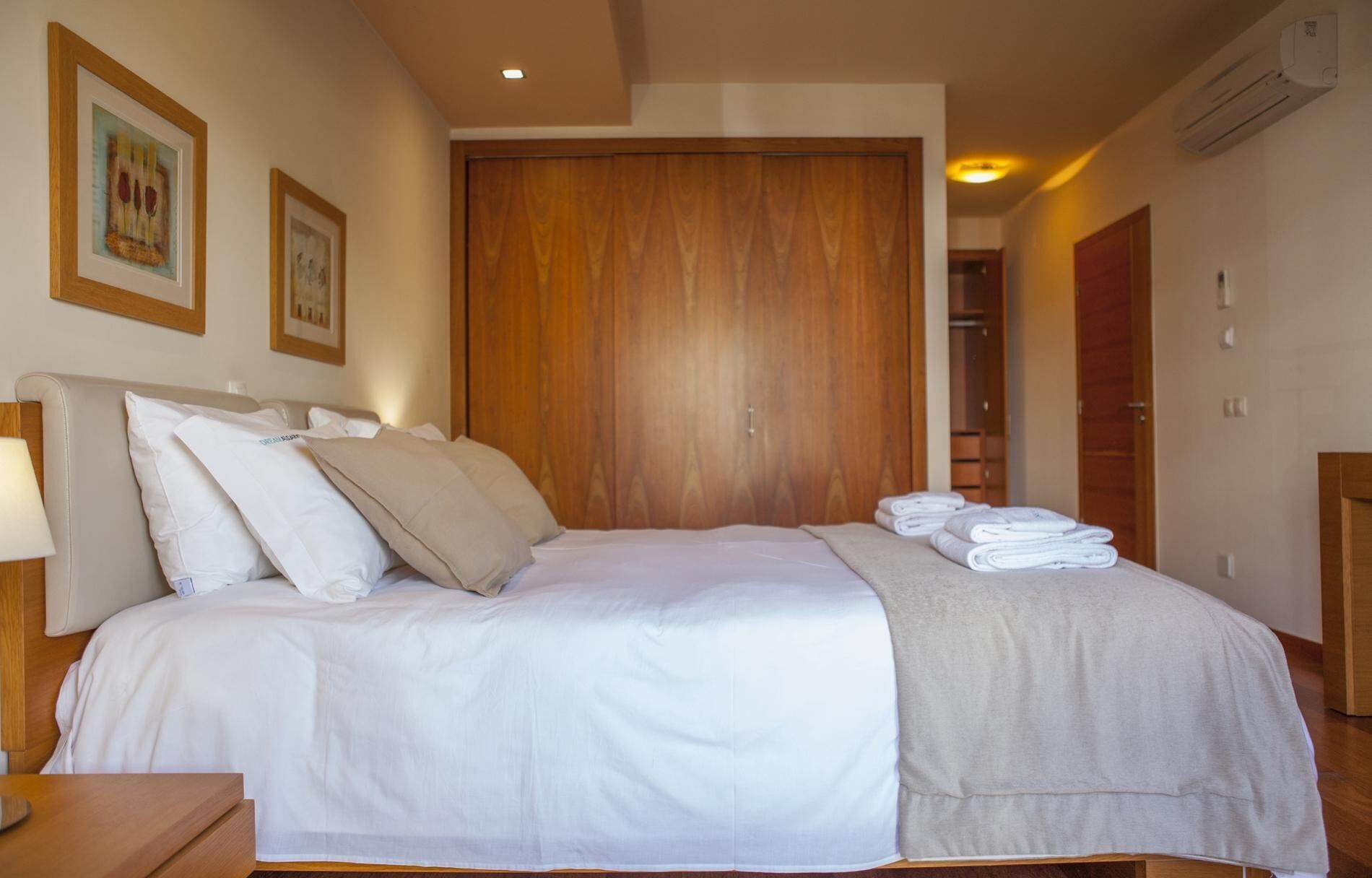 Una cama bien hecha con sábanas blancas y una manta gris, un armario de madera y una mesita de noche con una lámpara.