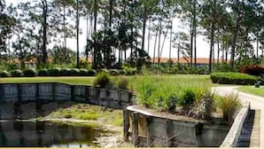 Um campo de golfe com uma ponte de madeira sobre um lago, rodeado por vegetação exuberante e árvores altas.