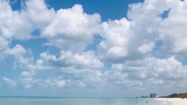 Uma praia com águas turquesa cristalinas, areia branca e nuvens dispersas.