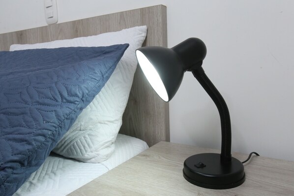 Una mesita de noche con una lámpara de escritorio negra, una cama con colcha azul y almohadas blancas, y un cabecero de madera.