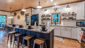 A modern kitchen with wooden cabinets, a large island with stools, pendant lighting, and a view through the window.