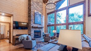 A cozy living room with a stone fireplace, wooden walls, and large windows offering a view of the outdoors.