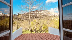 Une vue depuis une fenêtre donnant sur un toit recouvert de tuiles rouges, avec un arbre et une ville lointaine en contrebas.