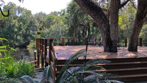 A wooden deck overlooking a river, surrounded by lush greenery and large trees. - River House - Overlooking the Scenic Withlacoochee River - Sleeps 4. (Inglis)