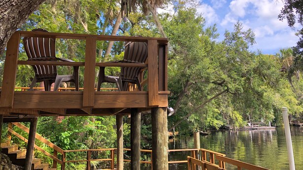 A wooden deck with two chairs and a table, overlooking a calm body of water with boats and lush greenery. - River House - Overlooking the Scenic Withlacoochee River - Sleeps 4. (Inglis)