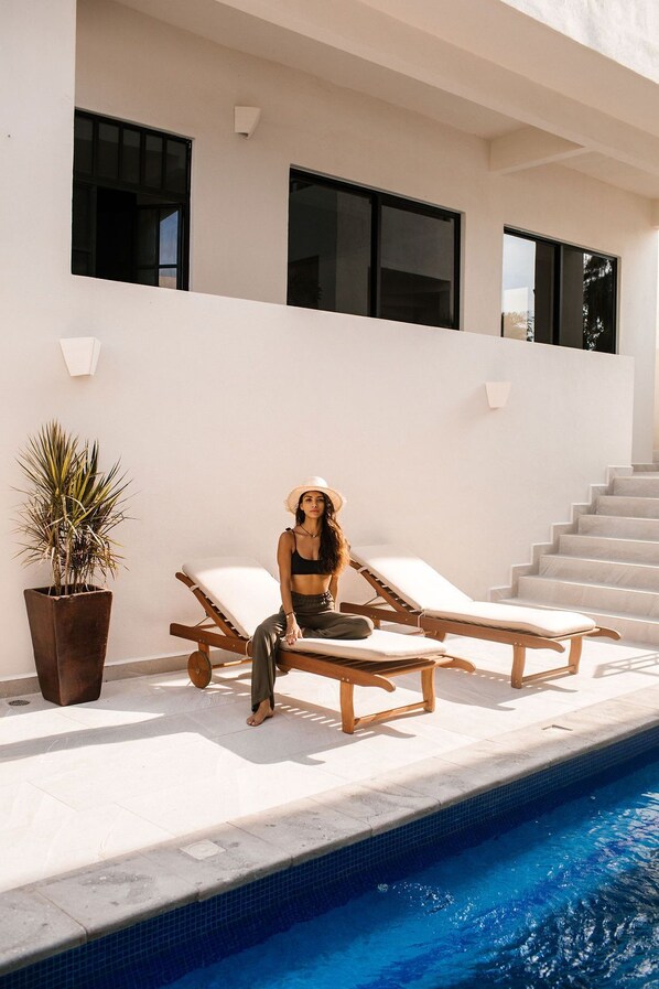 Un espace piscine extérieur moderne, avec une femme assise sur une chaise longue, une plante en pot et un escalier menant à un niveau supérieur.