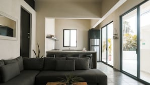 A modern living room with a dark sofa, a wooden coffee table, and a kitchen area with a large window. - Luxury Salwater Poolside Suite | Rooftop Terrace (Sayulita)