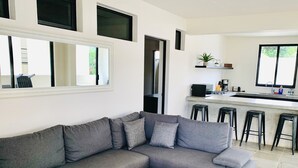 A modern living room with a grey sectional sofa, a wooden coffee table, and a kitchen area with a bar and stools.