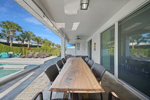 A covered outdoor dining area with a long wooden table and chairs, a pool, and a view of palm trees and a building. - Charming Oasis with Home Theater at Encore (Kissimmee)