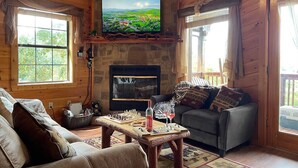 L’intérieur d’un chalet chaleureux est agrémenté d’une cheminée en pierre, d’un téléviseur fixé au-dessus, d’un canapé, d’une table basse avec un jeu d’échecs et des verres à vin, et d’une fenêtre avec des rideaux.