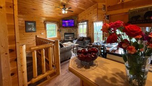 L’intérieur chaleureux d’un chalet avec des murs en bois, une cheminée, un coin repas comprenant une table et des chaises, et un téléviseur en arrière-plan.
