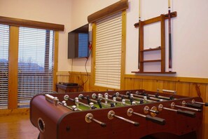 A foosball table in a room with wooden flooring, a television mounted on the wall, and windows with blinds.