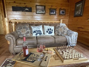 A cozy cabin interior with a wooden wall, a leather sofa with decorative pillows, a wooden coffee table with a bottle of wine, glasses, cookies, and a chessboard.