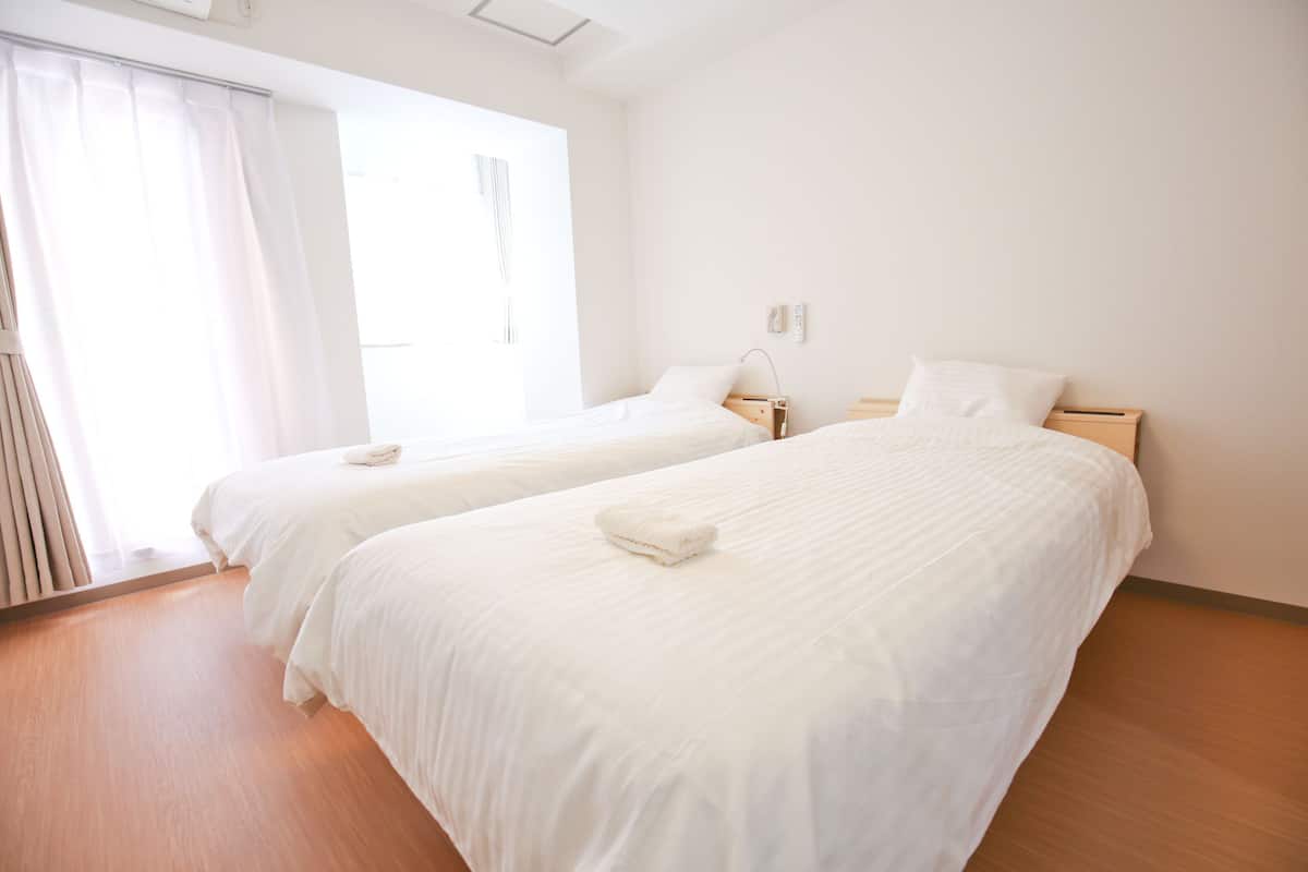 Habitación de hotel con dos camas individuales, suelo de madera, un ventanal grande con cortinas y una pared blanca.