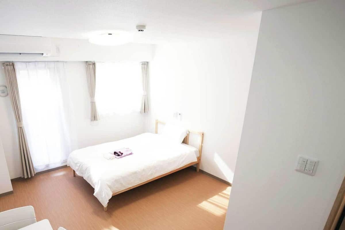Una habitación pequeña y limpia con una cama individual, una mesita de noche, una silla y una ventana con cortinas.