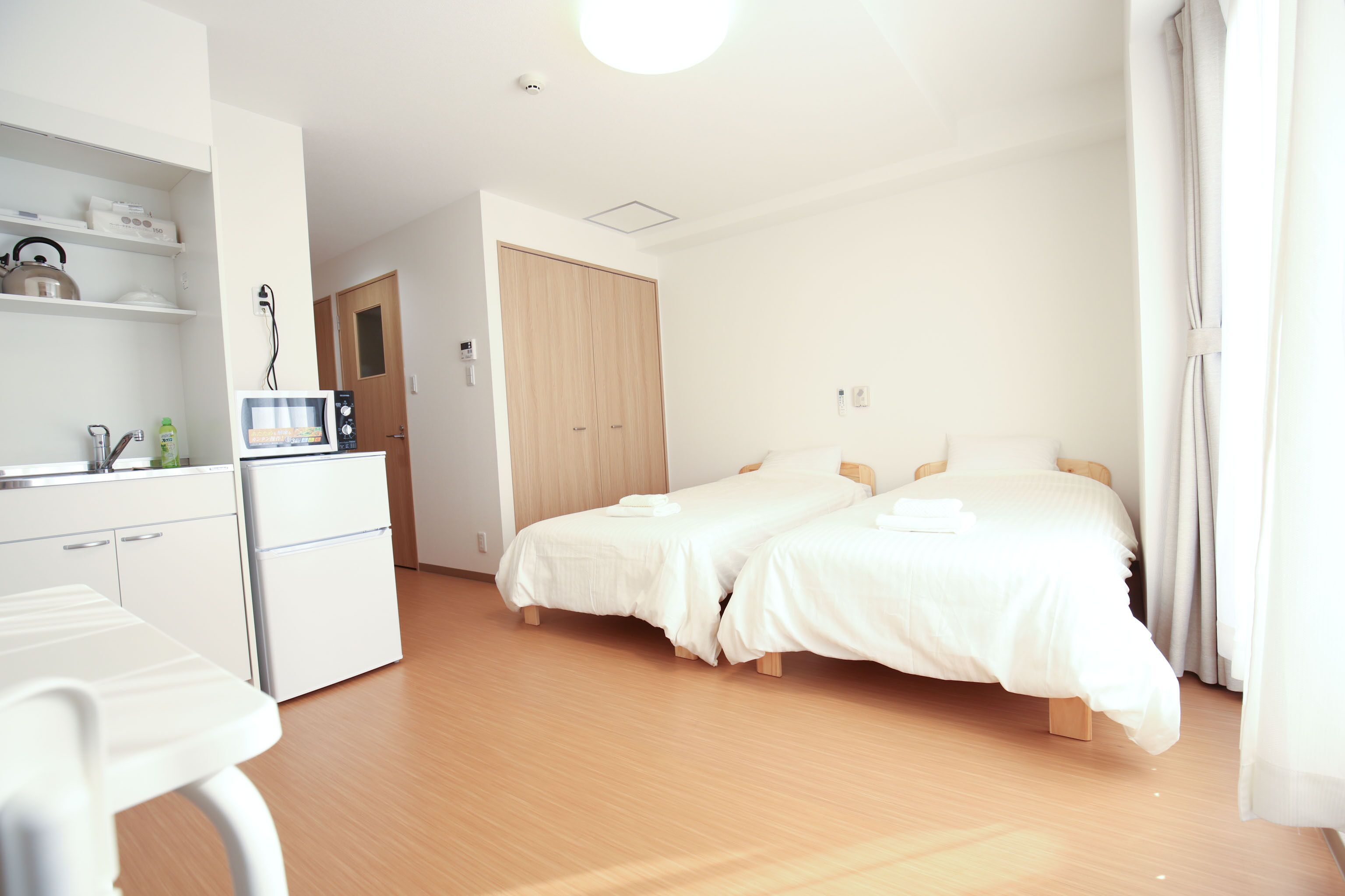 A hotel room with a kitchenette, two beds, a wooden wardrobe, and a bathroom visible through an open door.