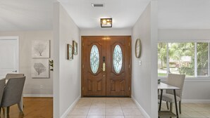 A clean, modern entryway with a wooden double door, a dining area with a table and chairs, and a window with blinds. - Beautiful Coastal-style Oasis! Heated POOL. 5min to BEACH! 8 Guests. (Seminole)