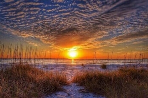 A sunset over a beach with a path leading to the water, surrounded by grass.