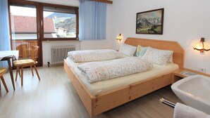 A bedroom with two beds, a wooden headboard, a window with a view of houses, a radiator, a chair, a table, and a bathtub. - Beautiful apartment for 4 guests with WIFI, TV, terrace and panoramic view (Fügen)