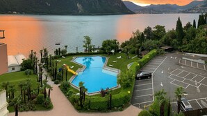 Ein Schwimmbecken mit Rutsche, umgeben von Grünpflanzen und einem Parkplatz, mit Bergen im Hintergrund.