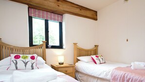 A bedroom with two beds, a window with a checkered valance, a bedside table with a lamp, and a wooden ceiling beam. - 3 bedroom accommodation in St Issey, Wadebridge (St Issey, Wadebridge)