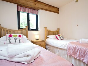 A bedroom with two beds, a window with a checkered valance, a bedside table with a lamp, and a wooden ceiling beam. - 3 bedroom accommodation in St Issey, Wadebridge (St Issey, Wadebridge)