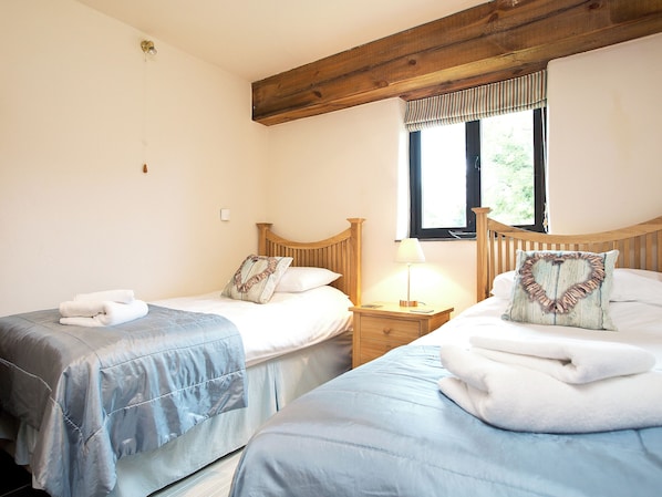 A bedroom with two beds, a nightstand with a lamp, a window with blinds, and a wooden ceiling beam. - 3 bedroom accommodation in St Issey, Wadebridge (St Issey, Wadebridge)