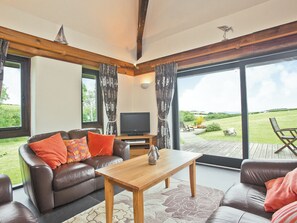 A living room with a sofa, a coffee table, a television, and a view of a grassy area through large windows. - 3 bedroom accommodation in St Issey, Wadebridge (St Issey, Wadebridge)