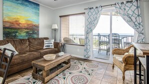 A living room with a brown leather sofa, a wooden coffee table, patterned floor tiles, a painting of a sunset, a balcony with wicker furniture, and a ceiling fan.