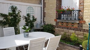 Een witte tafel met stoelen buiten, met een potplant, een metalen gieter en een bakstenen muur met een raam en bloembak.