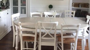 Een eetkamer met een witte tafel en stoelen, een kroonluchter, een schilderij van een brug en een boekenplank met boeken en decoratieve objecten.