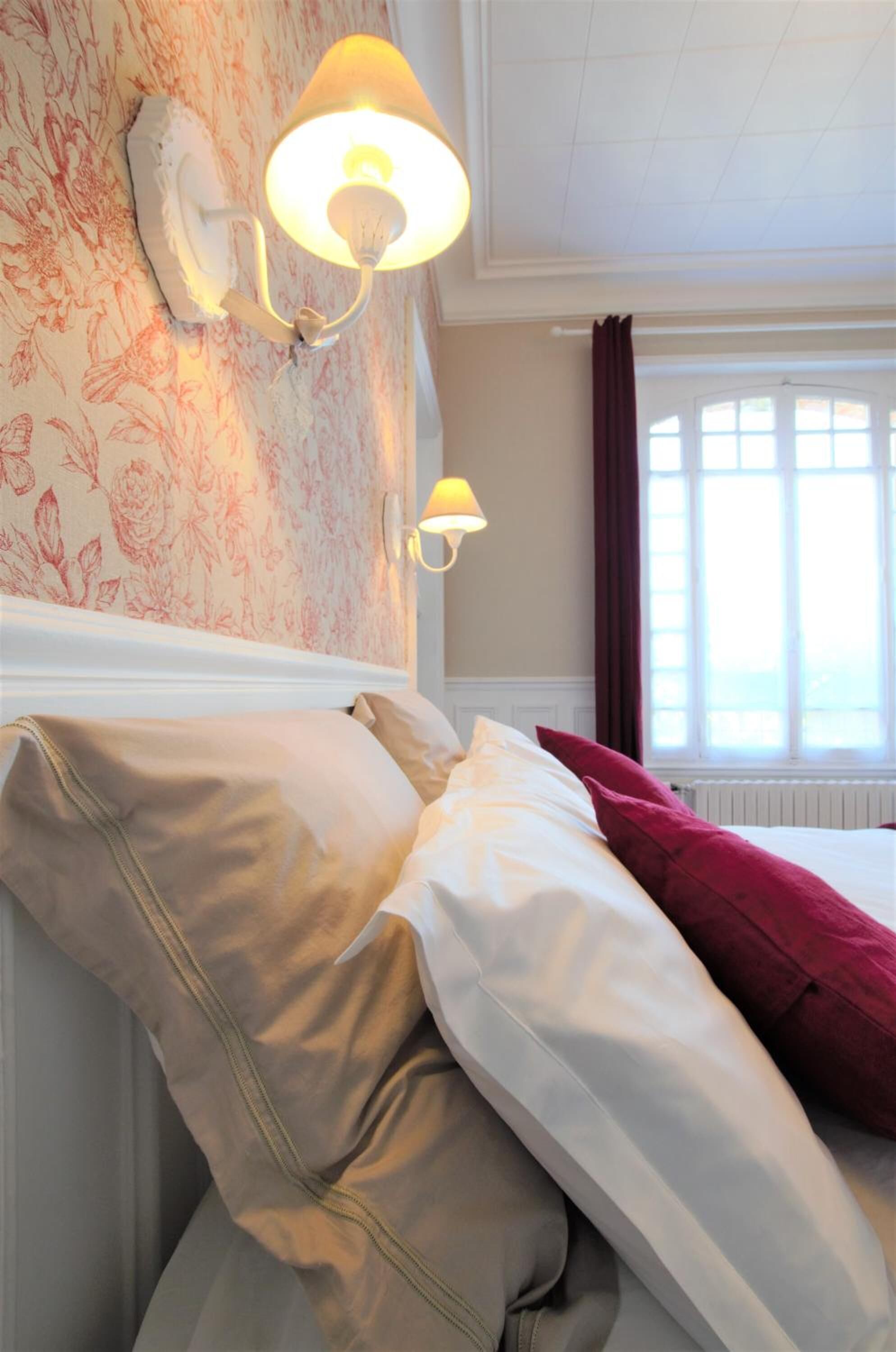 Una habitación de hotel con papel tapiz floral, dos lámparas de pared, una cama con almohadas blancas y rojas, y una ventana con cortinas.