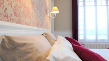 Una habitación de hotel con papel tapiz floral, dos lámparas de pared, una cama con almohadas blancas y rojas, y una ventana con cortinas.