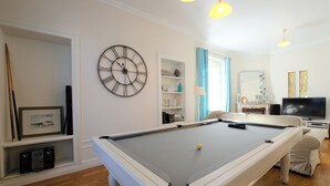 Een pooltafel in een goed verlichte kamer, met een klok aan de muur, een televisie en een boekenkast.
