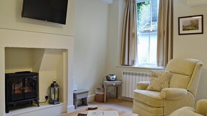 A living room with a fireplace, a television mounted on the wall, a comfortable armchair, and a window with curtains. - 2 bedroom accommodation in Keswick (Keswick)