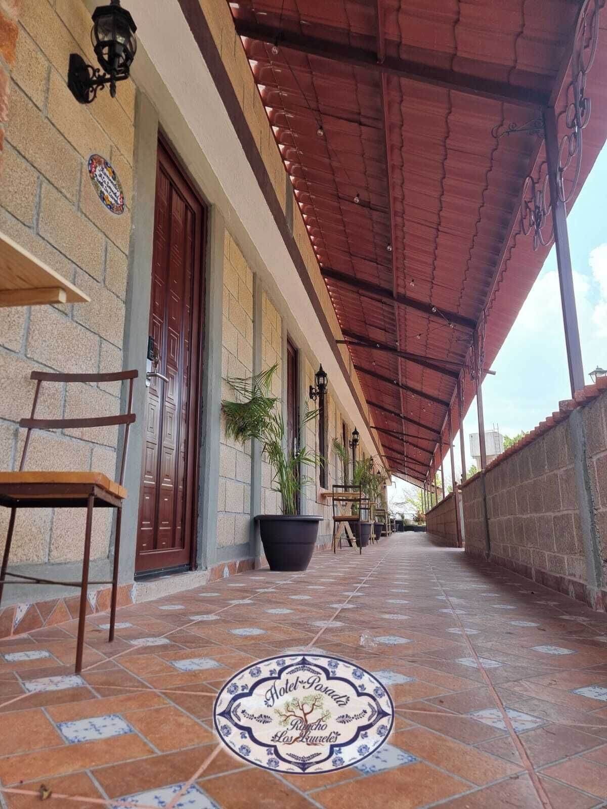 Uma galeria coberta com piso de azulejos, plantas em vasos e cadeiras de madeira.