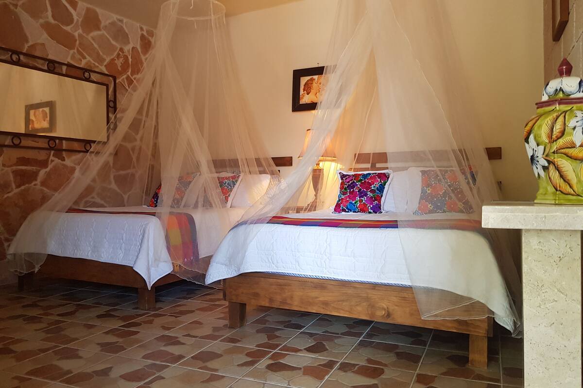 Quarto com duas camas, cada uma com colcha e almofadas. O quarto possui uma parede de pedra e piso de azulejos.