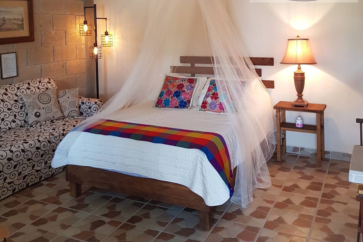 Quarto com cama sob dossel, mesa de cabeceira com luminária, relógio fixado na parede e quadro emoldurado.