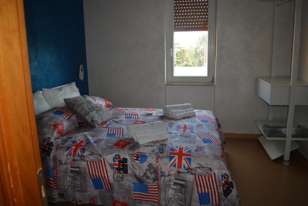 A bedroom with a bed covered in a flag-themed blanket, a window with blinds, and a white shelving unit.