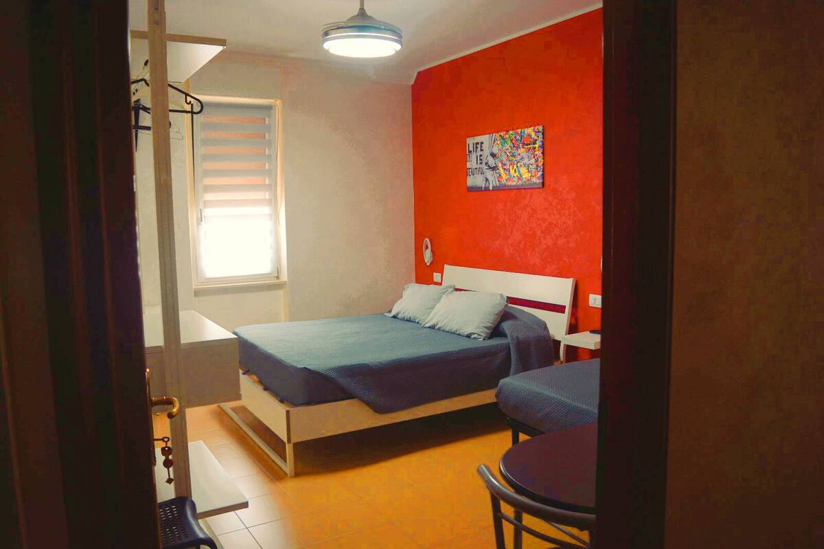 Un dormitorio con cama, una silla, una mesa, un ventanal con persianas y un cuadro en la pared.