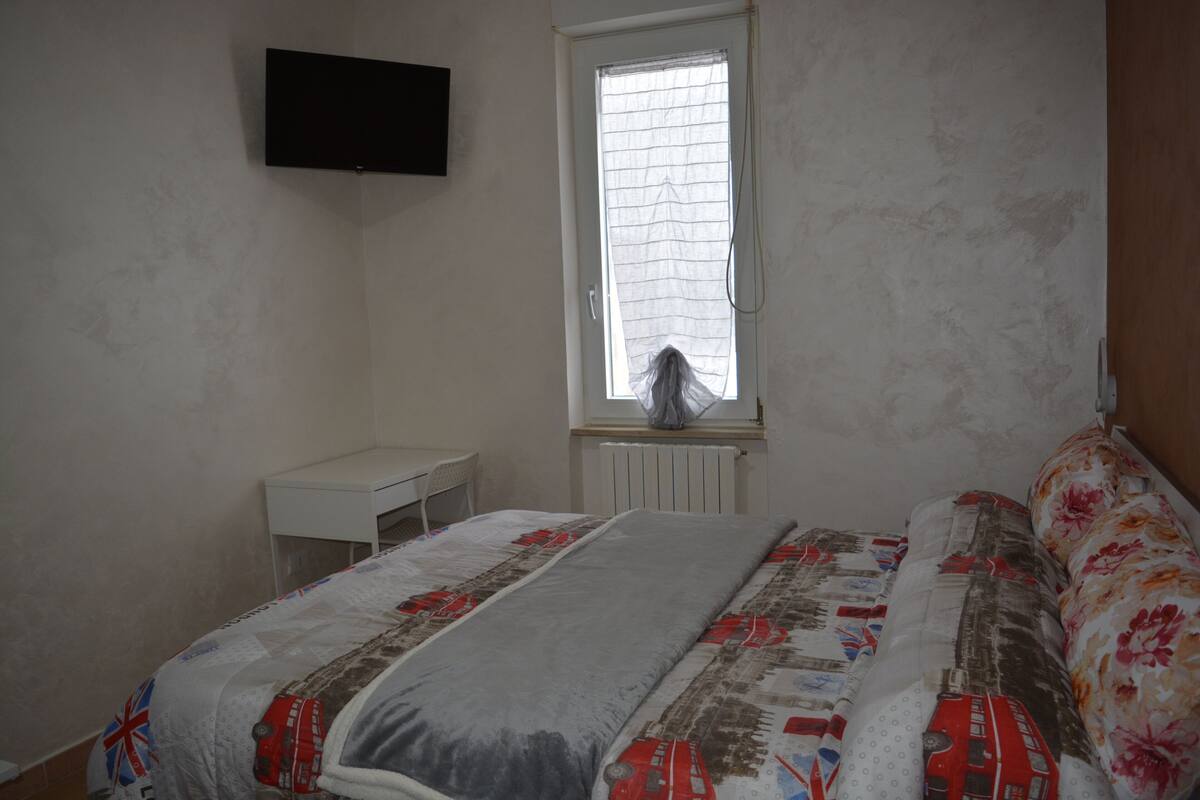 Un dormitorio con cama, ventana con persianas, mesita de noche blanca y televisión de pantalla plana montada en la pared.
