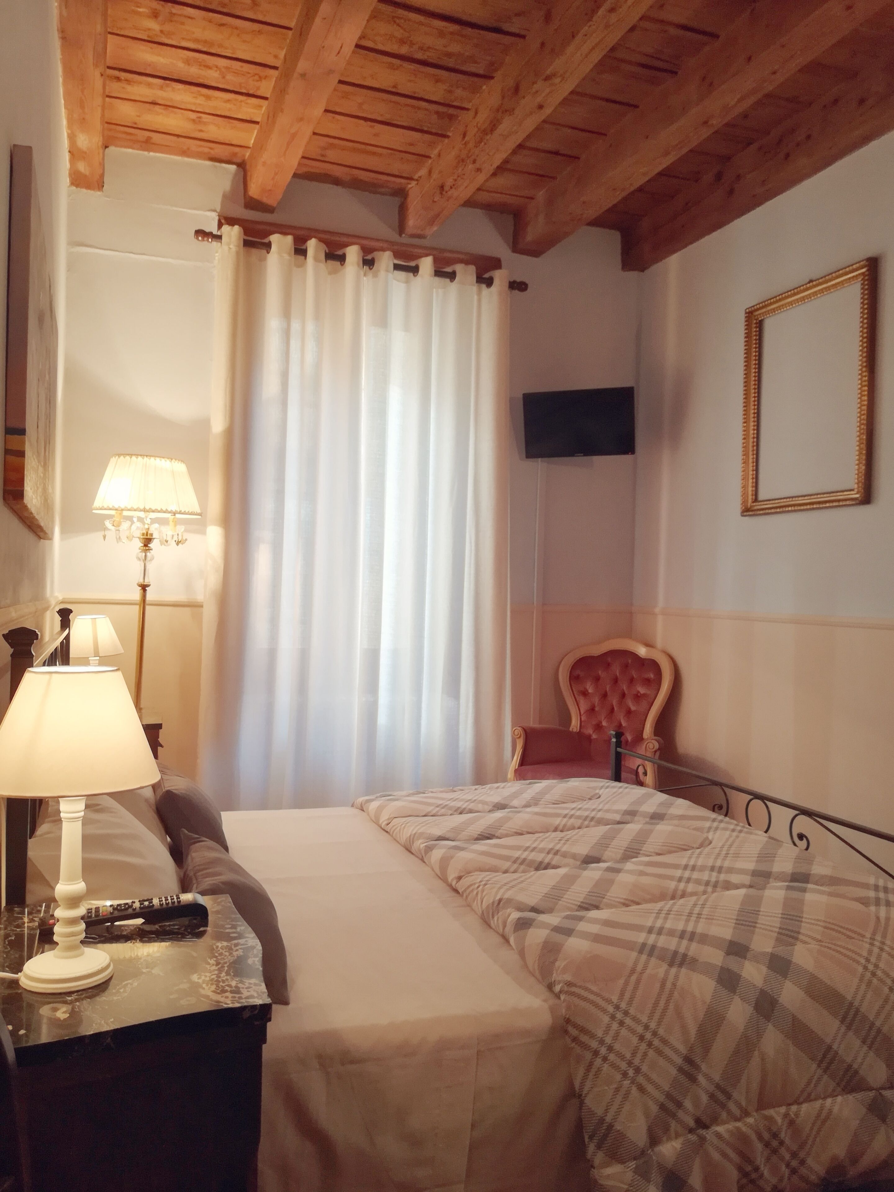 Habitación de hotel con cama, una silla, escritorio con lámpara, televisor y ventana con cortinas.