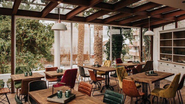 An outdoor dining area with wooden tables and chairs, large windows, and a view of trees and buildings. - 1930 Boutique Hotel (Arzua)